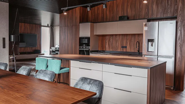 Moderná kuchynská linka v luxusnom interiéri, dominujúca elegantným dizajnom s kombináciou tmavého orechového dreva a matných béžových povrchov. V popredí sa nachádza veľký kuchynský ostrov so zásuvkami, ktorý slúži ako praktická pracovná plocha aj miesto na sedenie. Naľavo je umiestnený jedálenský stôl z masívneho dreva s rôznofarebnými čalúnenými stoličkami – niektoré sú v sivom, iné v tyrkysovom odtieni. V pozadí je vidieť vstavané spotrebiče vrátane rúry a mikrovlnky, ako aj americká chladnička s dávkovačom vody a ľadu. Steny a skrinky sú vyhotovené z materiálov s prirodzenou kresbou dreva, čo dodáva miestnosti teplo a štýlový vzhľad. Podhľad je riešený tmavými lamelami s bodovým osvetlením, ktoré rovnomerne nasvetľuje celý priestor a umocňuje moderný charakter kuchyne. Celkový dojem podporuje kombinácia útulnosti, sofistikovanosti a praktického využitia priestoru.